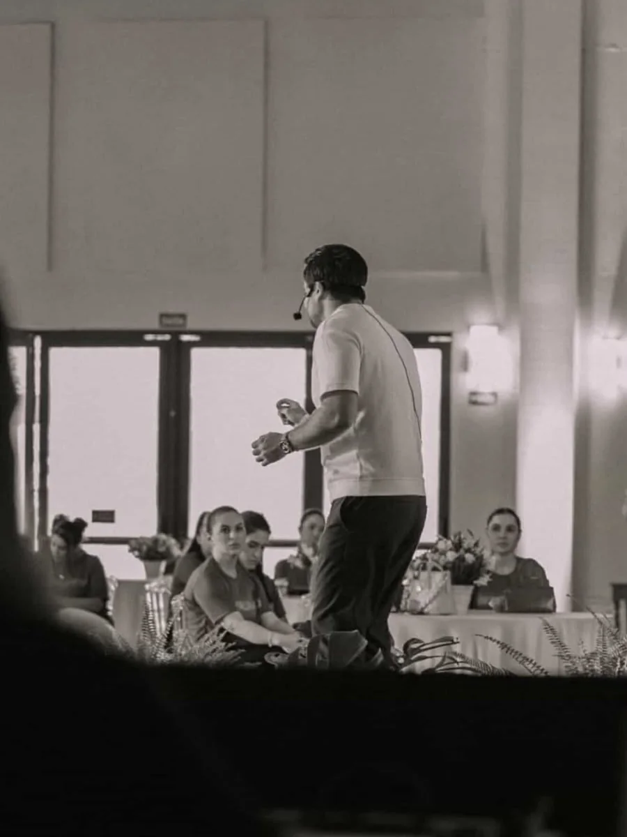 Minoru Raphael de costas, com microfone de cabeça, falando para a plateia, em foto em preto e branco
