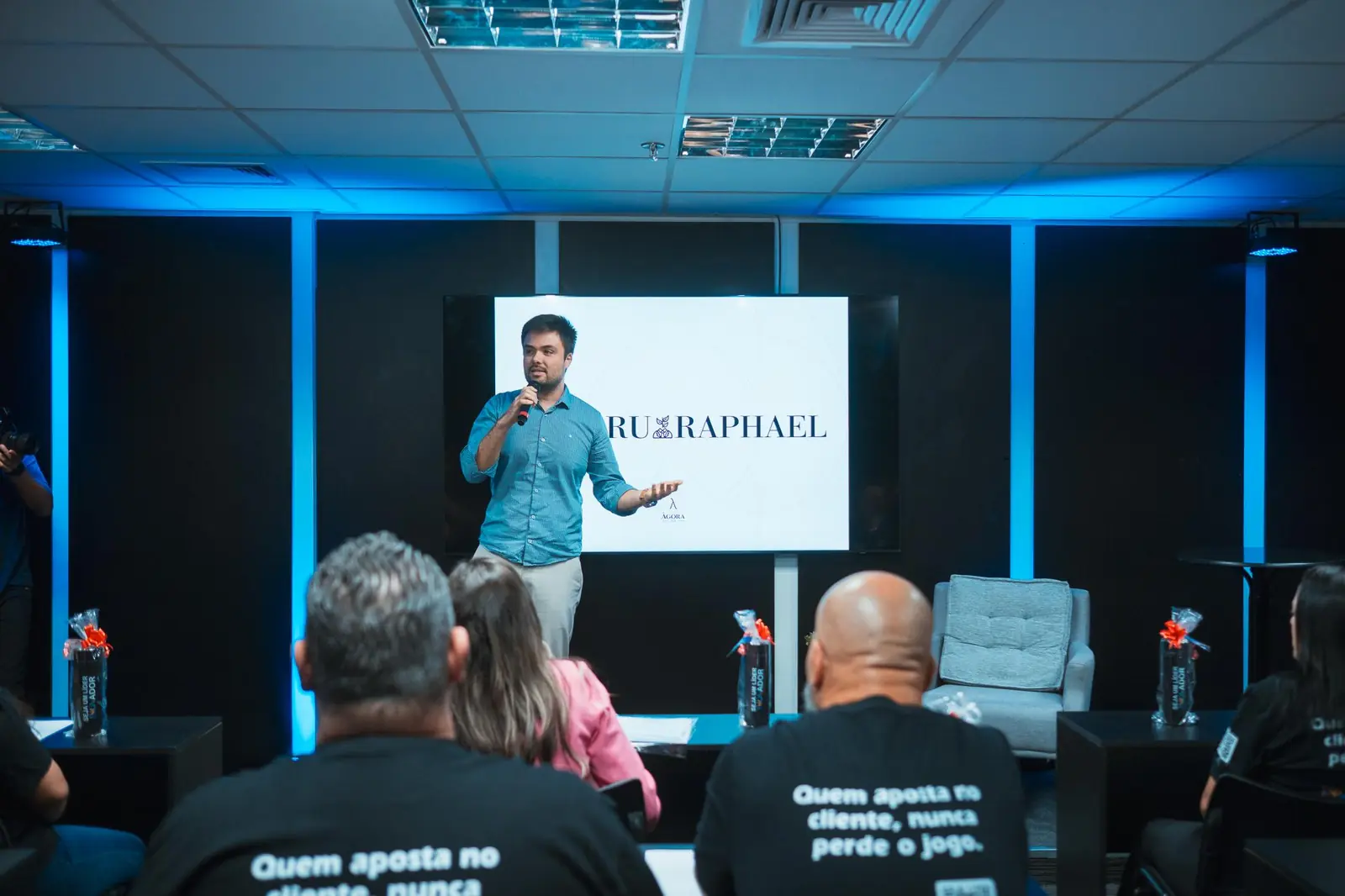 Minoru Raphael de camisa azul e microfone na mão, palestrando diante de um telão, com a plateia de costas em primeiro plano