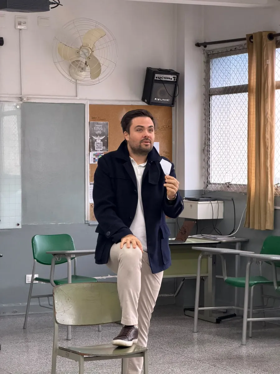 Minoru Raphael de blazer azul-marinho e camisa polo branca, com um pé apoiado numa cadeira, explicando algo a uma turma numa sala de aula de escola pública