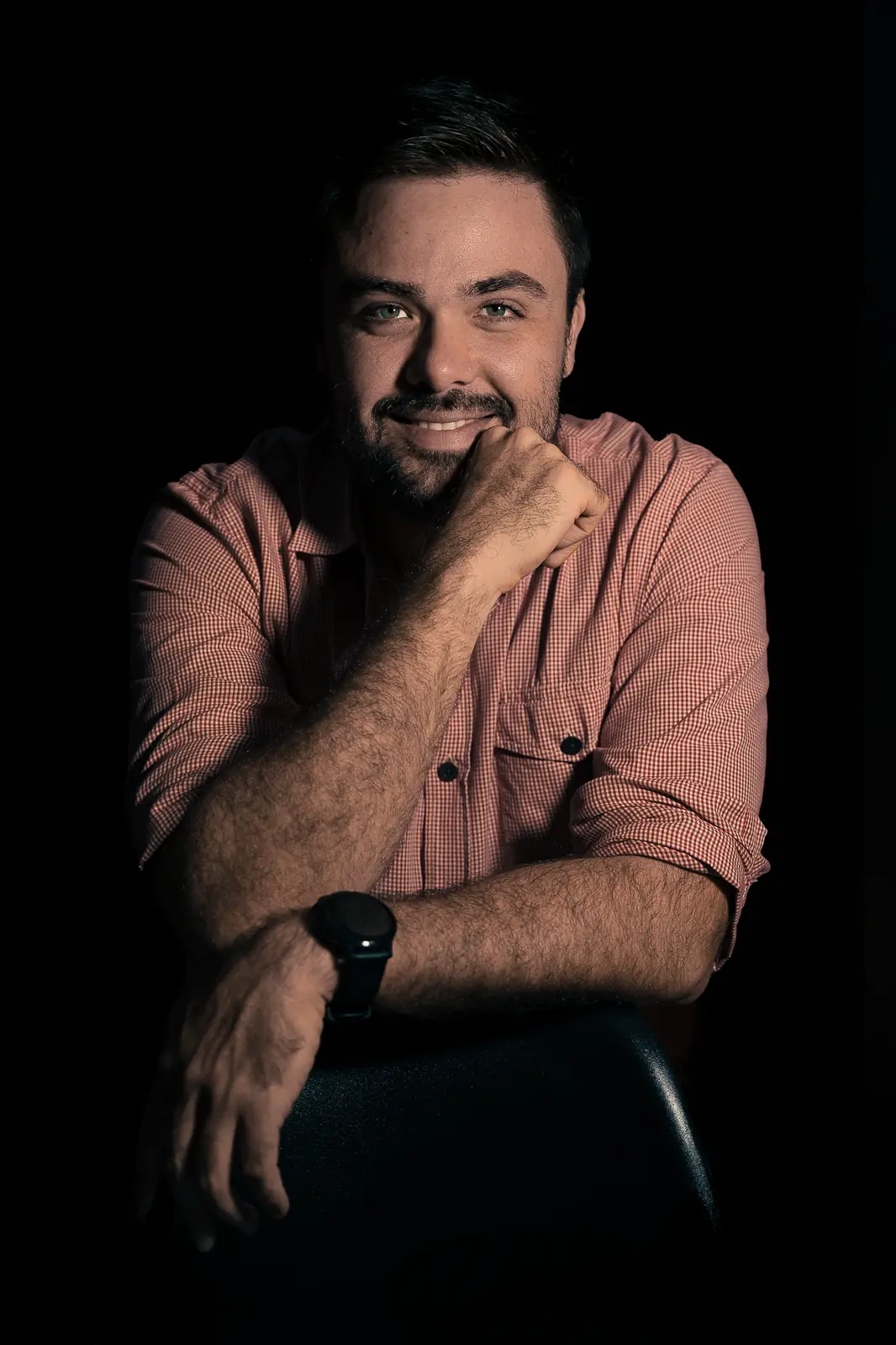 Minoru Raphael sorrindo, com o queixo apoiado na mão, de camisa xadrez, em retrato de fundo preto
