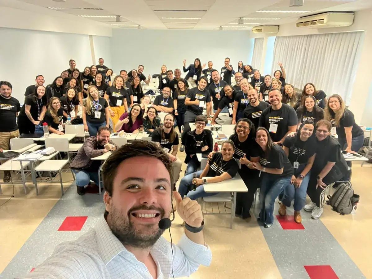 Minoru Raphael de camisa branca e microfone de cabeça, num selfie com uma turma de treinamento sorrindo, de camiseta preta, numa sala de aula