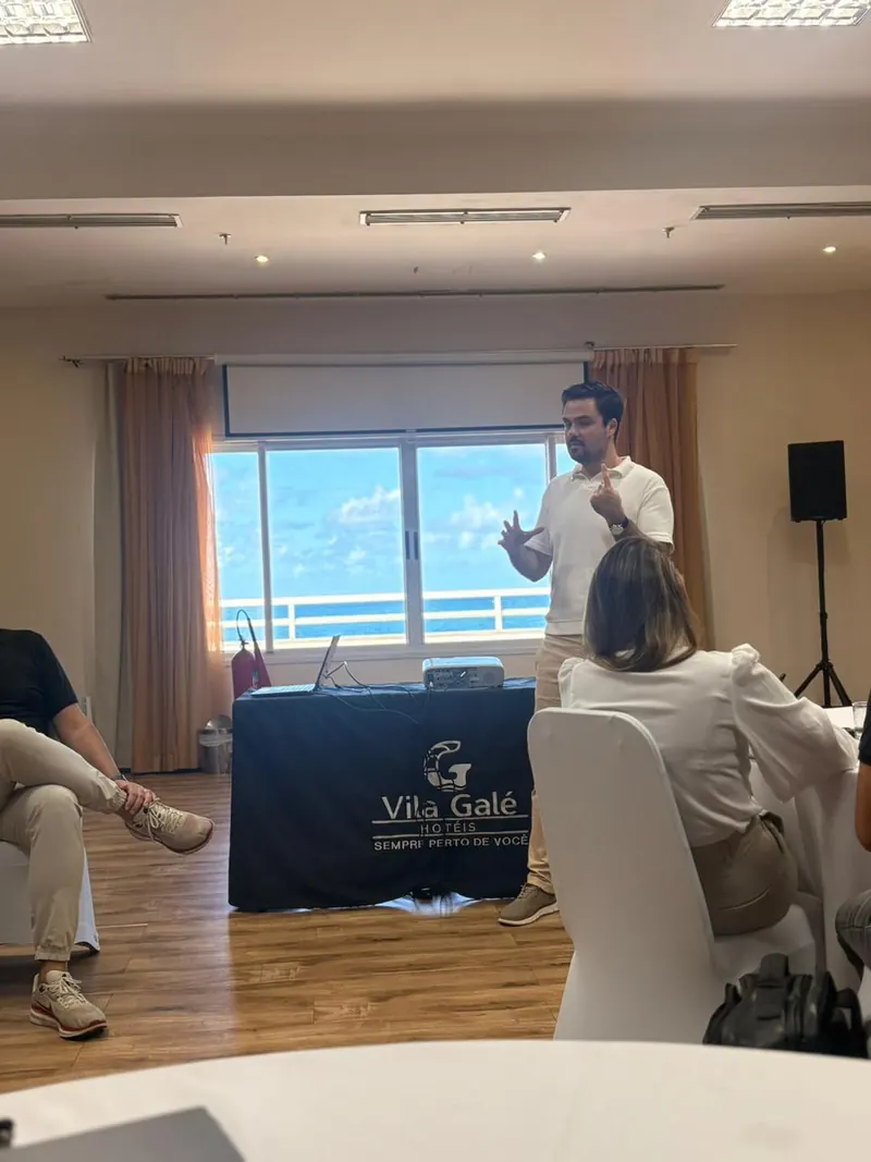 Minoru Raphael de camisa polo branca, gesticulando, durante uma palestra numa sala com janela para o mar