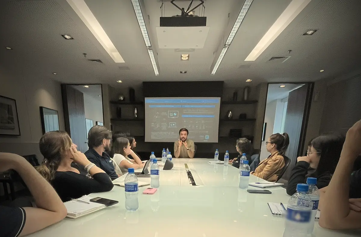 Minoru Raphael falando na ponta de uma mesa de reunião, diante de um telão, com a equipe sentada ao redor